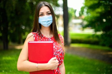 Güzel bir kız öğrenci dışarıda kitap tutuyor ve maske takıyor, koronavirüs konsepti.