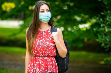 Parkta yürüyen ve kendini koronavirüsten korumak için maske takan bir kız öğrenci.