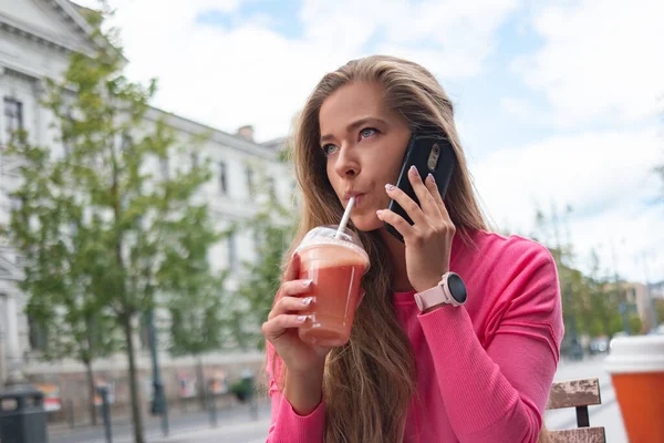 Woman using a straw to drink grapefruit juice outdoor in a city street while talking on the phone