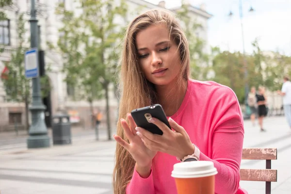 在城市里 一个年轻的女人在户外喝咖啡的时候用她的智能手机 — 图库照片