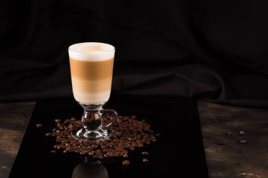 Siyah ayna arka planında kahve çekirdekleri olan bir bardak macciato latte. Kakaolu sütlü köpüklü kapuçino. Seçici odak, boşluğu kopyala