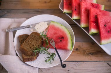 Yuvarlak beyaz tabak, bir dilim karpuz ve siyah ekmek, beyaz peynir ve biberiye, çatal ve bıçak, yan yana dikdörtgen beyaz karpuz tabağı.