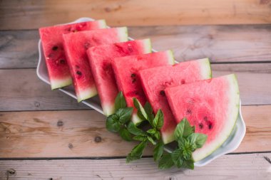 Tahta masanın üzerinde dikdörtgen beyaz bir tabak. İçinde karpuz dilimleri ve yeşil fesleğen yaprakları var.
