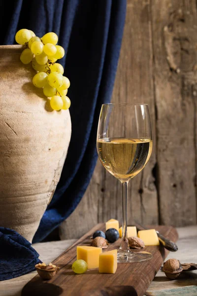White wine in glass goblet green yellow grapes, bottle and pottery jar, silver tray cheese platter wooden board, blue curtain with drapes, vintage old tavern still life, family winery, eco farm