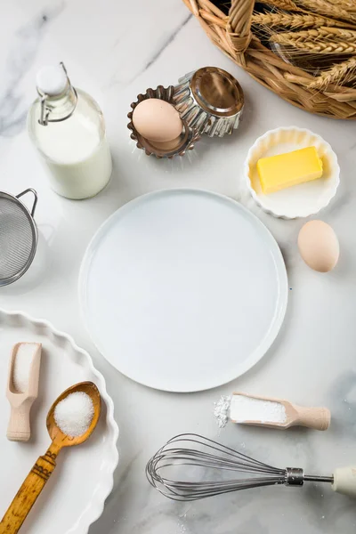 Ingredients for sweet baking on white marble background, eggs, milk jar, sugar, flour, butter, baking powder. Cookies or cake baking praparation. White round mock up for text in the center, copy space