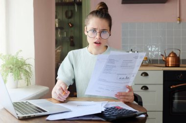 Faturalar elinde tutan mutfak masada oturan hayran genç kadın parers açılan ağzı ile seyir ifade, yüksek vergiler tarafından şaşırmış şaşkın. Mali sorunları kavramı