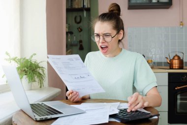 Faturalar elinde tutan mutfak masada oturan hayran genç kadın açık ağız, shouning ve srcreaming, parers arayan tarafından yüksek vergiler şaşırttı