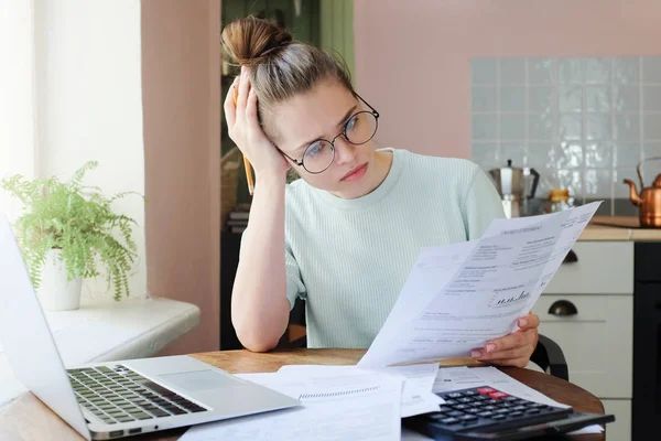 Young frustrated unhappy tired woman with financial troubles, sitting at kitchen table with papers, calculator and laptop computer, reading document