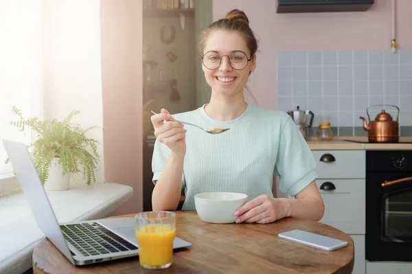 stock image Indoors shot of beautiful young smiling woman having delicious tasty breakfast, sitting at wooden table in modern colorful kitchen