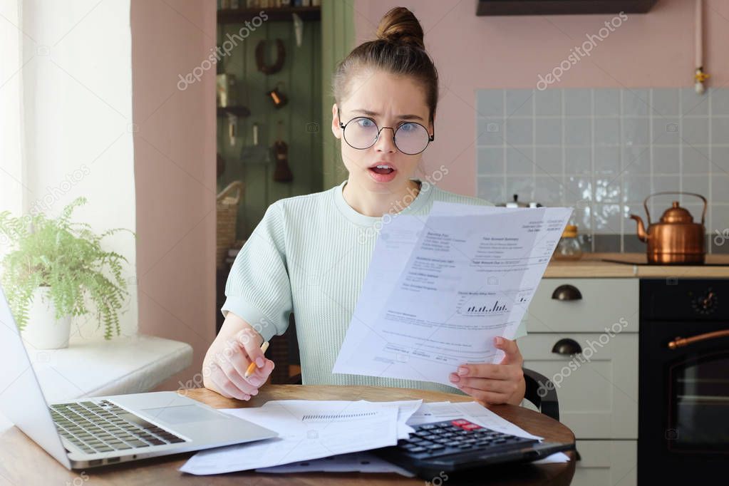 Amazed young woman sitting at kitchen table, holding utility bills in hand, looking at parers with opened mouth, puzzled expression, surprised by high taxes. Financial problems concept