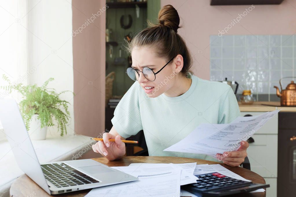 Amazed yong woman stressed over bills, sitting at kitchen table, looking at laptop screen in complete shock, astonished with amount of unpaid bills. People and finances concept