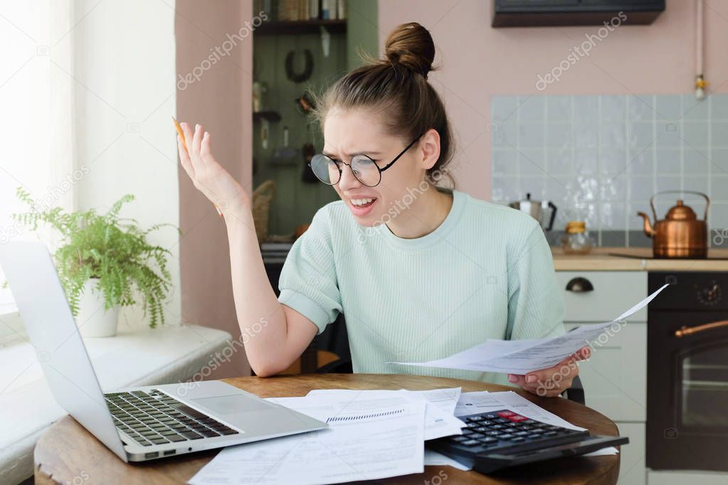 Indignant angry irritated young woman looking at utility bill, worried about troubles with mortgage payment to bank, having serious problems with debt, frustrated about high taxes, 