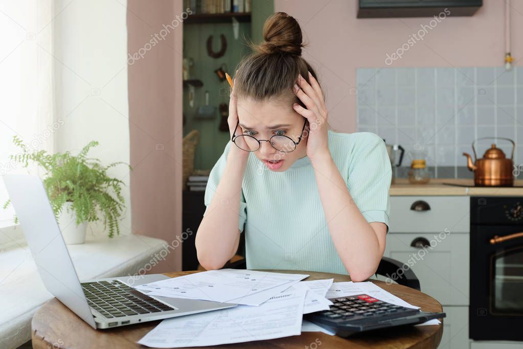 Young anxious worrying woman looks desperately to bills, faces problems with paying taxes, holding head with both hands