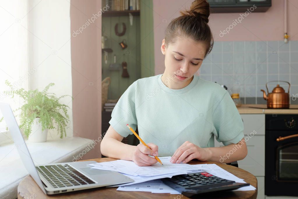 Portrait of young woman, sitting at kitchen table, filling application form, calculating expences, trying to solve budget problems, writting something on papers, managing with utility bills