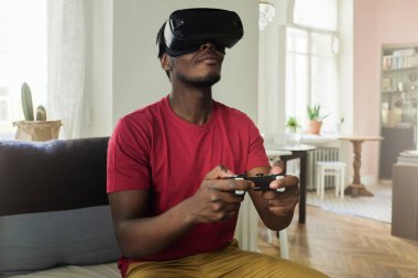 Afrika kökenli Amerikalı adam vr bardaklarda kanepede oturan iskambil video oyunları sanal gerçeklik kulaklık ile ve uzaktan kumanda ile oyun çubuğu 