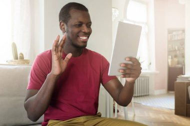 Gün ışığı atış Afrikalı-Amerikalı gencin ailesi veya kız arkadaşı, bir elinizle sallayarak ile online konuşmak tablet kullanarak rahat aydınlık salonda oturan 