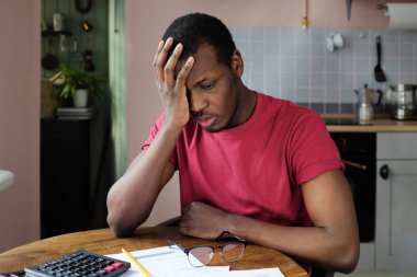 Hayal kırıklığına uğramış mutsuz yorgun Afro-Amerikan olan genç adam mali sıkıntılar, belgeler, hesap makinesi ve yüz palm jest ile mutfak masada oturan