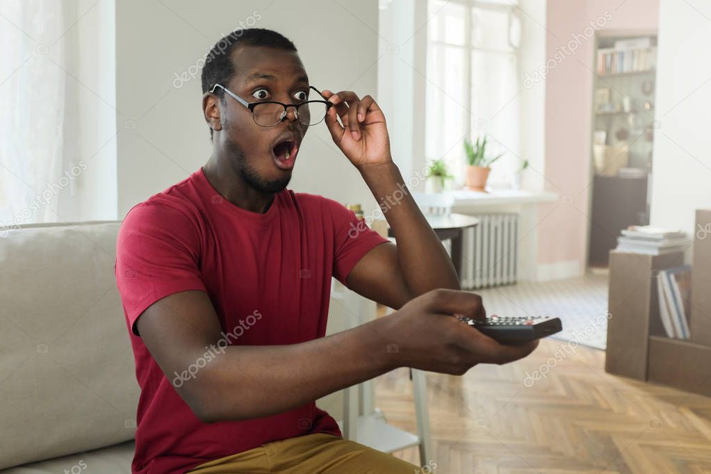 Young astonished dazed shocked amazed african american man rising up eyeglasses, sitting on couch, watching TV show with mouth open and holding remote control