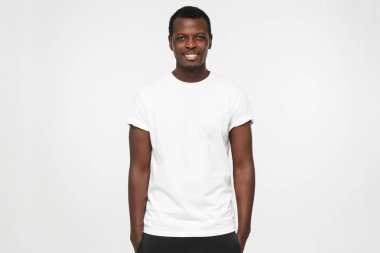 Elleri ceplerinde duran, logo veya metin, gri arka plan üzerinde izole kopya alanı olan boş beyaz t gömlek giymiş genç Afro-Amerikan adam