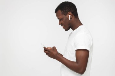 İleti değiş tokuşu veya Internet'te sörf yaparken akıllı telefon ekranına bakarak gri arka plan üzerinde izole boş Beyaz tişörtlü, yakışıklı Afrikalı-Amerikalı adam portresi yan