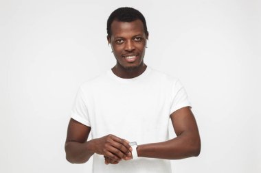 Gri arka plan üzerinde izole dokunmatik ekranlı akıllı tutan kablosuz kulaklık ve boş beyaz t tişört giyen genç gülümseyen Afrikalı-Amerikalı adam izliyor 