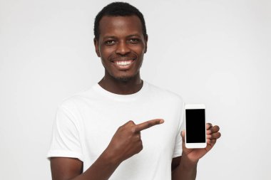 Akıllı telefon sunulması ve boş siyah perde ile boşaltmak reklamlar için parmağınızla işaret Afro-Amerikan yakışıklı beyaz t gömlek gri arka plan üzerinde izole