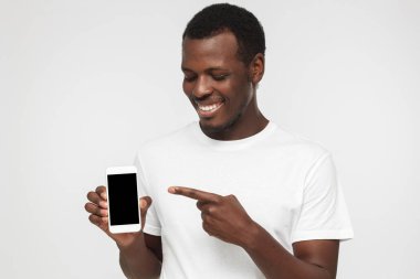 Boş telefon tutarak ve ekran gösteren ilginç bir teklif için işaret t gömlek giymiş genç Afro-Amerikan erkek gülümseyen yakışıklı beyaz zemin üzerine izole