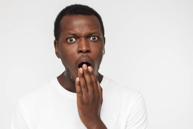 Close up photo of young african american man isolated on gray background dressed in blank white t shirt, covering mouth with hand, experiencing deep astonishment and fear clipart