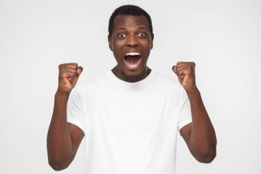 Yumruk ve ağız içinde neşeli öfori duygu sanki açıldı olması kenetlenmesi duygusal Afrikalı-Amerikalı adam boş beyaz t gömlek gri arka plan üzerinde izole mutluluk zafer hakkında