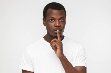 Young handsome African american man isolated on gray background wearing blank white t shirt and pressing index finger to lips as if asking other to keep silent about exciting secret clipart
