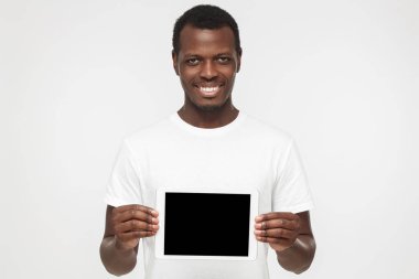 Pozitif Afrikalı-Amerikalı adam ayakta beyaz t gömlek holding tablet ve ürün veya hizmet danışmanlık sanki boş ekran mutlu gülümseme ile gösterilen gri arka plan üzerinde izole stüdyo resmi