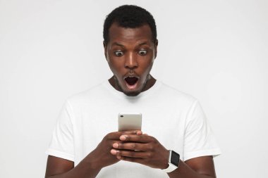 Ağzı açık boş beyaz t gömlek, yakışıklı genç Afro-Amerikan adam smartphone, sürpriz ve karışık görünümlü tutarak portresi kapatın