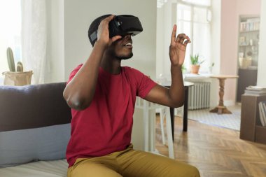 Gördüğü nesneye dokunun çalışılıyor sanki yukarı işaret rahat kıyafetler Vr kulaklık oyun, olanaklarının yaşandığı giyiyor Oturma odasında kanepede oturan genç Afrika adam fotoğrafı