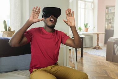 Genç korkmuş Afrikalı adamın üzerinde şaşırmış duygu oyun ortamında hareket sanki hareketleri el ile yapma sanal gerçeklik gözlüklü oturan kanepe evde boş zaman zaman harcama resmi