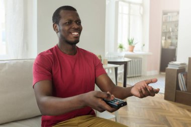 İç oturma odası izlerken Tv programları veya uzaktan kumanda ile haber oturan genç Afrika adam portresi elinde, sanki duygu şaşkın silah yaymak ve ifade bu konuda şüphe