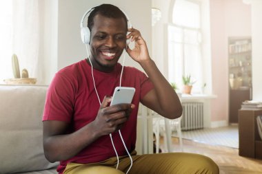 Afrika kökenli Amerikalı gencin evde boş zaman zaman harcama iç dikey smartphone kullanarak ve şarkı ve gülümseme ile tezahürat ekran bakarak büyük beyaz kulaklık üzerinden müzik