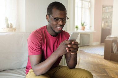 Kırmızı T-shirt ve el Internet tarama veya rahat gülümseme ile arkadaşı ile mesaj alışverişi smartphone ile kanepede oturan gözlük takan genç Afro-Amerikan adamın yatay resmi