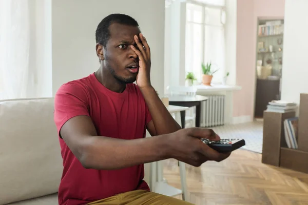 Indoor photo of handsome African American man holding remote control while watching television, pressing one palm to his face as if he sees nonsense on screen and does not agree with its content