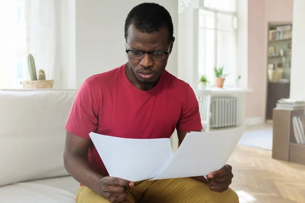 Interior photo of handsome African man dressed in casual clothes staying indoors, sitting, comparing numbers in sheets of bill or documents, feeling worried and facing great financial challenge