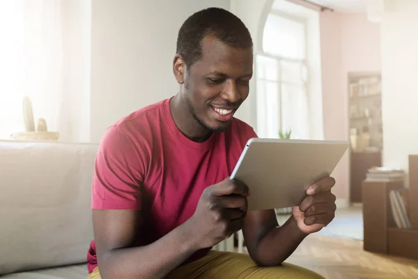 英俊的非洲裔美国人的室内图片坐在白天在他的房间里显示轻松的笑容 因为他看着他的平板电脑屏幕上握着手 浏览互联网或观看视频 — 图库照片