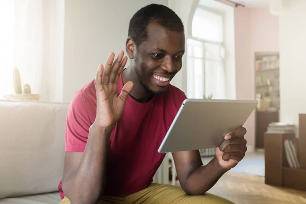 Indoor portrait of young African American man spending afternoon in living room holding tablet waving friendly to speaker on screen, saying hello, smiling, feeling relaxed and sharing positive mood