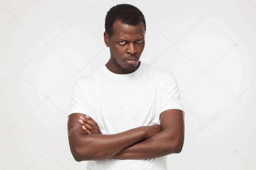 Horizontal shot of young african american man in blank white t shirt with arms crossed isolated on gray background, looking at camera with anger