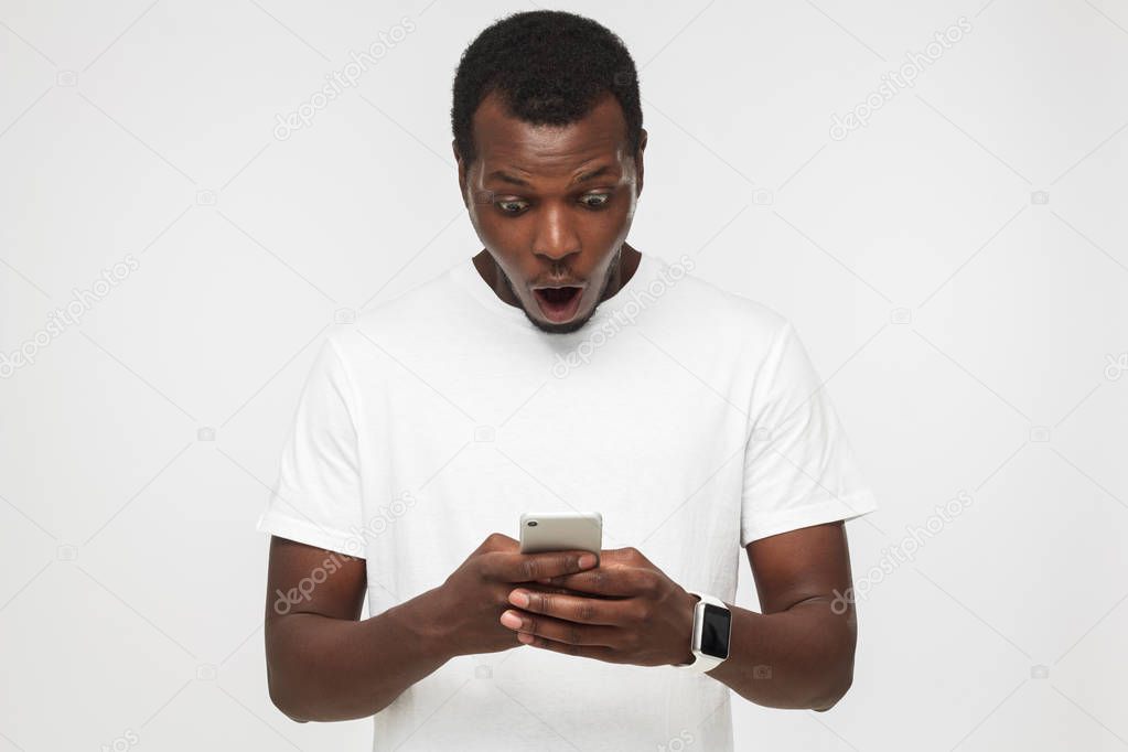 Handsome African American man isolated on white background looking to screen of smartphone with mouth round because of deep astonishment and shock of great news