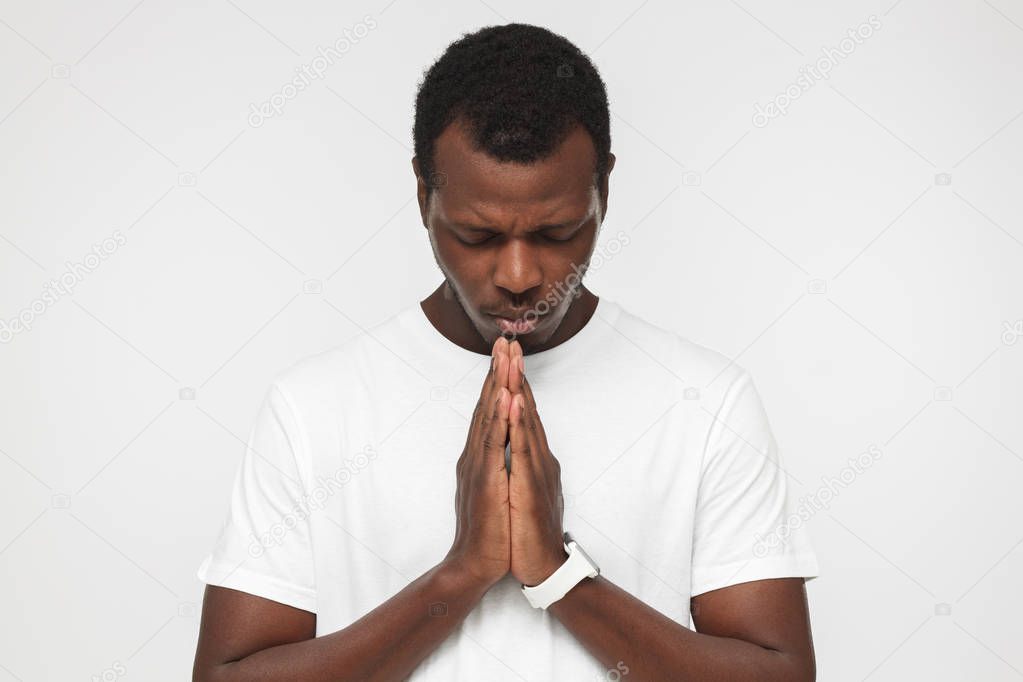 Closeup of young african american man isolated on gray background looking stressed; putting hands together as if he is praying with closed eyes to overcome depression