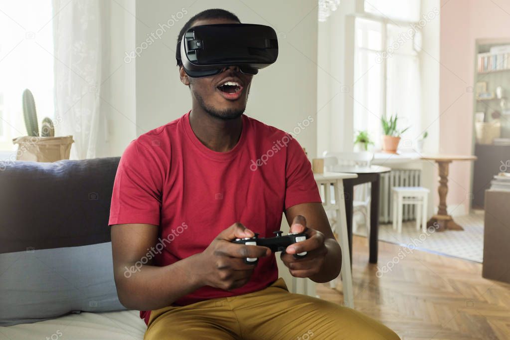 Indoor picture of African man dressed in red T-shirt having rest on sofa with virtual reality set on and gaming console in hands looking astonished with what he sees on screen, feeling positive