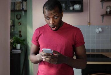 Yakışıklı Afrika adam cep telefonu tamamen sürpriz olağanüstü başarı veya zafer yaşıyor ekranda gördüğü ile her iki elinde tutan onun mutfakta ayakta closeup portresi