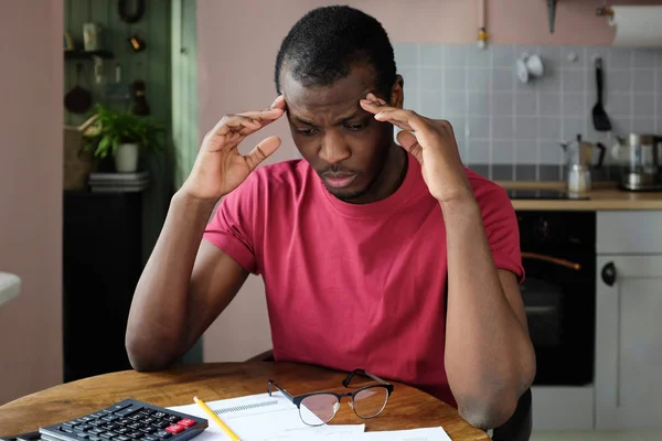 Indoor shot of young African American man pressing fingers to temples feeling pain in his head trying to figure out accounting details of his bills, looking helpless and uneasy, not knowing what to do