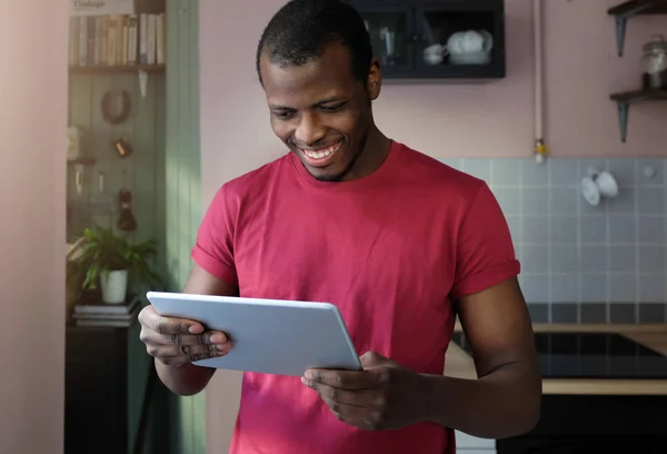 年轻的非洲裔美国人的室内图片享受闲暇时间在家里穿着休闲服装手持平板电脑在手看满意的网页内容与积极的微笑 — 图库照片