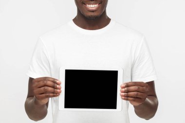 Pozitif Afrikalı-Amerikalı adam ayakta beyaz t gömlek holding tablet ve ürün veya hizmet danışmanlık sanki boş ekran mutlu gülümseme ile gösterilen gri arka plan üzerinde izole stüdyo resmi