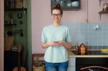 Closeup photo of smiling European female staying at home alone standing in her kitchen communicating happily via cellphone looking straight at camera feeling confident, relaxed and satisfied with life clipart